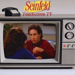 Può includere: Un televisore in stile vintage mostra una scena con una persona in camicia rossa. Sopra lo schermo, il testo recita Seinfeld Touchscreen TV, accompagnato da un piccolo modello di telefono nero. Il televisore presenta due grandi manopole.