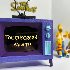 May include: A purple mini TV toy with the text "TOUCHSCREEN MINI TV" on the screen. The TV has light blue knobs and a matching antenna. In the background, there are figurines of characters from The Simpsons.