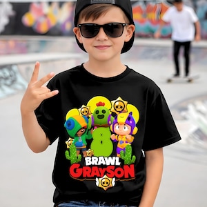 May include: A black t-shirt featuring a colorful graphic of cartoon characters, the text "Brawl Grayson", and a star logo. The person in the image is wearing a black cap and sunglasses.