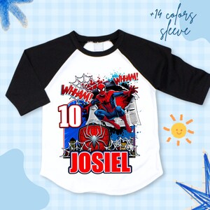 Pu&ograve; includere: T-shirt bianca e nera con maniche a raglan con grafica di Spider-Man. La grafica include il numero 10, il nome "JOSIEL" e la parola "WHAM!". La maglietta ha uno sfondo blu e bianco con un disegno di sole e stella.
