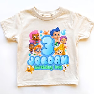 May include: A cream-colored short-sleeve t-shirt featuring a graphic print of Bubble Guppies characters, a large blue number 3, and the text JORDAN birthday boy in blue and green bubble-style lettering, surrounded by starfish and bubbles.