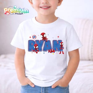 Może przedstawiać: Biała koszulka z napisem "RYAN" w kolorze niebieskim, obramowanym na czerwono, z grafiką Spider-Mana. Koszulka ma okrągły dekolt i kr&oacute;tkie rękawy. Tekst "Premium Printtees" znajduje się w lewym g&oacute;rnym rogu.