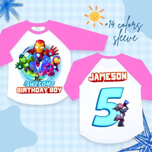 Puede incluir: Camiseta blanca y rosa de estilo ragl&aacute;n con un gr&aacute;fico de superh&eacute;roes y el texto "THE AWESOME BIRTHDAY BOY". La parte trasera de la camiseta muestra el nombre "JAMESON" y el n&uacute;mero "5".