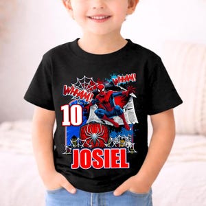Pu&ograve; includere: T-shirt nera con grafica colorata di Spider-Man. Il design include il numero 10, il nome JOSIEL e la parola "WHAM!" in stile fumetto. La maglietta ha scollo tondo e maniche corte.