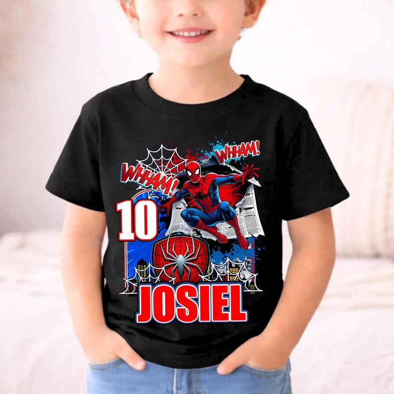 Pu&ograve; includere: T-shirt nera con grafica colorata di Spider-Man. Il design include il numero 10, il nome JOSIEL e la parola "WHAM!" in stile fumetto. La maglietta ha scollo tondo e maniche corte.