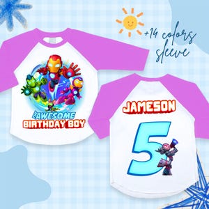 Puede incluir: Camiseta ragl&aacute;n blanca y morada con un gr&aacute;fico de superh&eacute;roes y el texto "THE AWESOME BIRTHDAY BOY". La parte trasera de la camiseta muestra el nombre "JAMESON" y el n&uacute;mero "5" en azul.