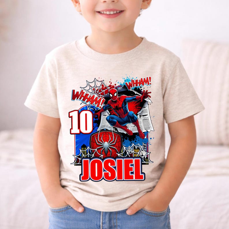 Pu&ograve; includere: T-shirt beige con grafica colorata di Spider-Man. Il design include il numero "10", il nome "JOSIEL" ed effetti sonori da fumetto. Il supereroe &egrave; raffigurato in una posa dinamica, con un costume rosso e blu.