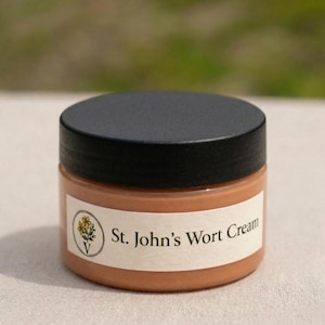May include: A small jar of St John's Wort Cream with a black lid. The jar has a white label with the product name and a floral illustration. The cream inside is a light brown colour.