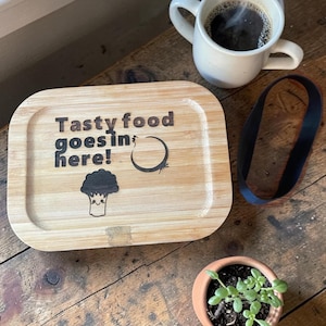 May include: A wooden lunch box with the text "Tasty food goes in here!" and a broccoli illustration. A white mug with coffee and steam, a black band, and a small potted plant sit on a wooden surface.