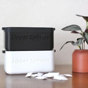 May include: A black and a white rectangular container stacked on top of each other, each with the text "Lipper Spitters" embossed on the front. A small terracotta pot holds a plant with dark green and purple leaves. Small white paper pieces are scattered in front.