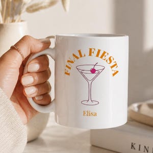 Puede incluir: Taza de cerámica blanca con las palabras "FINAL FIESTA" en naranja y una ilustración de copa de cóctel en morado. La taza también tiene el nombre "Elisa". La taza está sostenida por una persona.