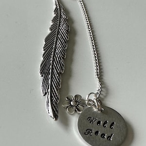Peut inclure: Marque-page en métal argenté avec un motif de plume, un petit charme de fleur et un pendentif circulaire. Le pendentif porte l'inscription "Well Read". Le marque-page est conçu pour être inséré dans un livre.