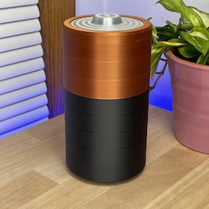May include: A large, cylindrical object resembling a battery. The top section is copper-colored, while the lower section is black. The top has a silver-colored, circular cap. The object is sitting on a wooden surface.
