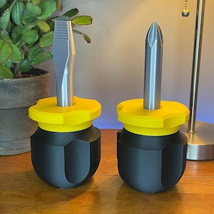 May include: Two screwdrivers with black handles, yellow tops, and silver metal shafts. One has a flathead tip, and the other has a Phillips head tip. The tools are displayed on a wooden surface.