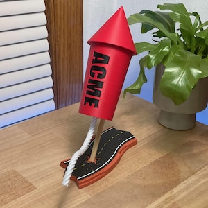 May include: A red cylindrical rocket model with a conical top and the word ACME printed in black letters. The rocket is mounted on a wooden stick attached to a base shaped like a section of black asphalt road with yellow dashed lines.