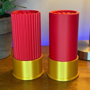 以下が含まれることがあります： 赤と金の装飾的な散弾銃の薬莢型の容器が2つ。1つは垂直リブのディテールがあり、もう1つは滑らかです。どちらも金色のベースがあり、木製の表面に置かれています。容器は円筒形で、鮮やかな赤色をしています。