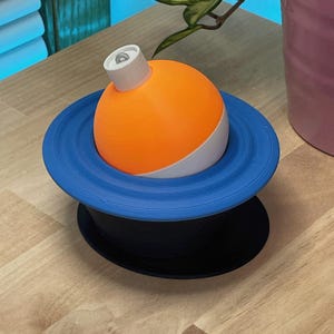 May include: A 3D-printed decorative object featuring a bright orange sphere with a white and blue base, resembling a planet. The sphere has a white top and sits on a blue ring, which is on a black base. The design is modern and geometric.
