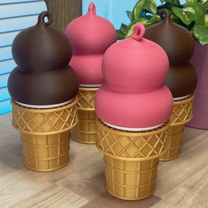 May include: Four decorative ice cream cone-shaped containers. Two have brown tops, and two have pink tops. The tops have a swirl design. The bases are gold-colored waffle cones. The containers are on a light brown surface.