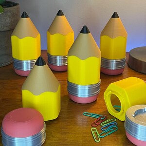 May include: A collection of pencil-shaped containers in yellow, tan, and black. The containers have a silver band around the middle and a pink base. Several containers are upright, while others are open, revealing the interior. Paper clips are scattered nearby.