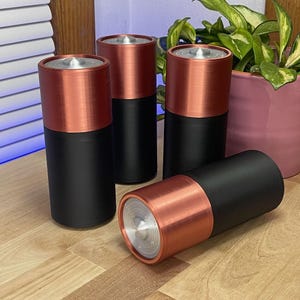 May include: Four cylindrical objects with a copper-colored top and a black base are displayed. One object is lying on its side, while the others stand upright. The objects have a metallic appearance and a modern design. The background includes a wooden surface and a plant.