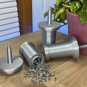 May include: Silver-colored push pin holders with a cylindrical design and a conical top. One holder is open, spilling out a pile of silver push pins. The holders are on a wooden surface.