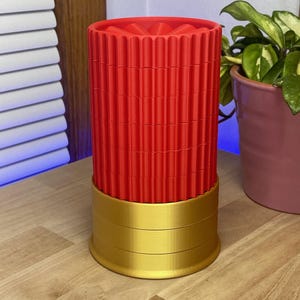 Op de afbeelding: Een rood en goudkleurige decoratieve container. De cilindrische container heeft een geribbelde rode bovenkant en een goudkleurige basis. De container staat op een houten oppervlak. Een plant in een pot is zichtbaar op de achtergrond.