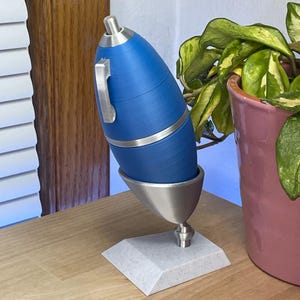 May include: A blue and silver pen with a unique design. The pen has a cylindrical body with a silver band and a silver clip. It is displayed on a silver stand with a gray base. A pink flower pot with green leaves is in the background.