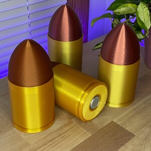 May include: Four decorative bullet-shaped objects. The tops are a metallic copper color, and the bases are a bright gold. One bullet is lying on its side, revealing a silver-colored interior. The objects are arranged on a wooden surface.