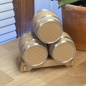 May include: A set of three light tan plastic barrels stacked in a pyramid formation on a matching tan base. Each barrel features horizontal molded lines and a silver-colored circular rim at the front end, creating a uniform and structured appearance.