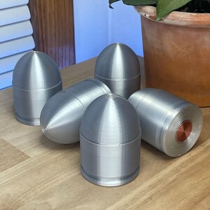 May include: A collection of four silver-colored, bullet-shaped objects with rounded tops and cylindrical bases. Each piece features a smooth, metallic finish and a distinct copper-colored circular insert visible on the flat base of one unit.