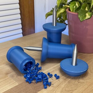 May include: Blue push pin holders with silver metal pins. One holder is tipped over, spilling blue push pins onto a wooden surface. The holders are cylindrical with a wide base and a silver metal pin. A pink pot with a green plant is in the background.
