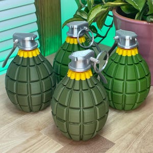 Può includere: Quattro repliche di granate decorative verde oliva con dettagli gialli e argentati. Le granate hanno una superficie strutturata e un anello di trazione. Gli oggetti sono disposti su una superficie di legno.