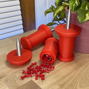 May include: Red push pin holders with silver metal spikes. Several holders are shown, some upright and some on their sides. A pile of red push pins are scattered on a wooden surface.