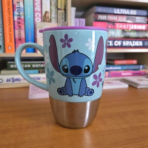 Op de afbeelding: Lichtblauwe reismok met het personage Stitch uit Lilo & Stitch. De mok heeft een roestvrijstalen basis, een lichtblauw handvat en een paarse deksel. Stitch wordt afgebeeld in een zittende positie, omringd door witte en paarse bloemen.