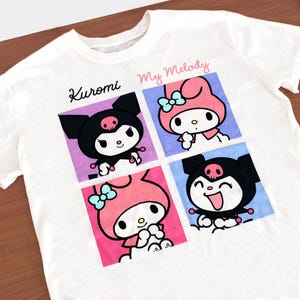 May include: White t-shirt featuring four cartoon character squares. The top left square says "Kuromi" and the top right says "My Melody". The characters are in black, pink, and blue. The shirt is made of a soft material.