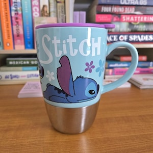 Op de afbeelding: Lichtblauwe reismok met een roestvrijstalen basis, een paarse deksel en een handvat. De mok toont het personage Stitch uit Lilo & Stitch, met het woord "Stitch" in het wit. Bloemenaccenten sieren de mok, ideaal voor warme of koude dranken.