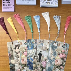 May include: A collection of decorative bookmarks with colorful tassels. The bookmarks feature vintage-style designs with images of roses, butterflies, and the Eiffel Tower. Accompanying the bookmarks are small cards with text that reads "Your order made my day" and "Be the best version of you."