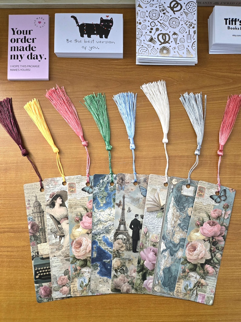 May include: A collection of decorative bookmarks with colorful tassels. The bookmarks feature vintage-style designs with images of roses, butterflies, and the Eiffel Tower. Accompanying the bookmarks are small cards with text that reads "Your order made my day" and "Be the best version of you."