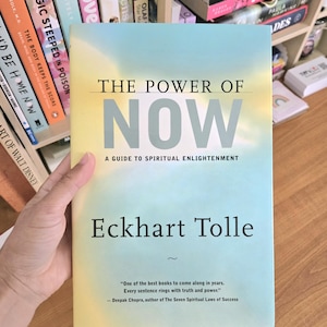 May include: A book titled "The Power of Now" by Eckhart Tolle. The cover has a gradient of yellow and blue with the title in large, bold letters. The subtitle reads "A Guide to Spiritual Enlightenment."