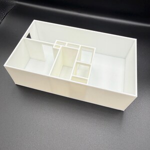 Op de afbeelding: Een witte rechthoekige organizer met meerdere compartimenten van verschillende afmetingen. De organizer is gemaakt van een solide, ondoorzichtig materiaal en is ontworpen om kleine items in op te bergen. De compartimenten worden gescheiden door dunne wanden.