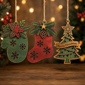 May include: Three hanging wooden ornaments: a green mitten with snowflake cutouts and pinecone accents, a red stocking with snowflake cutouts and pinecone accents, and a green Christmas tree with heart cutouts and the text Merry Christmas.