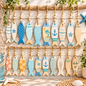 Op de afbeelding: Een collectie van twintig houten visornamenten die aan twee horizontale stukken drijfhout hangen. Elke vis heeft unieke geschilderde ontwerpen, waaronder ankers, palmbomen, zeesterren, bloemmotieven en strepen, geaccentueerd met natuurlijke houten kralen.