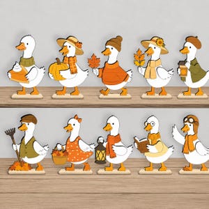 Puede incluir: Una colección de diez figuras de patos de madera plana dispuestas en dos estantes. Cada pato blanco tiene pico y patas naranjas, vistiendo accesorios otoñales como bufandas, gorros, suéteres y sosteniendo objetos como calabazas, hojas y libros.