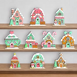 Peut inclure: Une collection de onze ornements décoratifs en bois plat en forme de maisons en pain d'épices et d'une tasse. Chaque pièce présente des couleurs pastel, notamment rose, vert menthe et marron, avec des détails blancs imitant le glaçage.