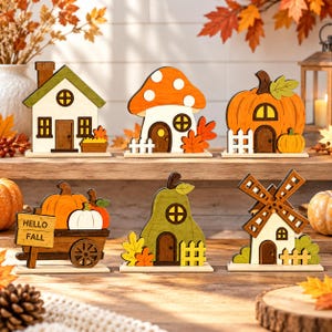 Op de afbeelding: Een set van zes houten tafeldecoraties met een huis, een paddenstoel, een pompoen, een wagen met pompoenen en een bordje met de tekst HELLO FALL, een peervormig huis en een windmolen, allemaal beschilderd in herfstkleuren met witte hekjes.