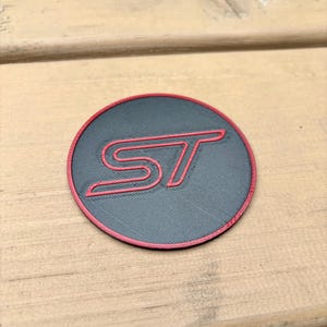 Peut inclure: Un emblème circulaire avec un fond noir, une bordure rouge et les lettres rouges ST en relief au centre. La surface de l'emblème présente une finition texturée et superposée, caractéristique d'une fabrication par impression 3D.