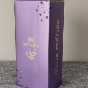 May include: A rectangular, lavender-colored box with the Stanley logo and the initials "OR" in white. The box is decorated with star shapes in various colors and the name "olivia rodrigo" is printed vertically on the side.