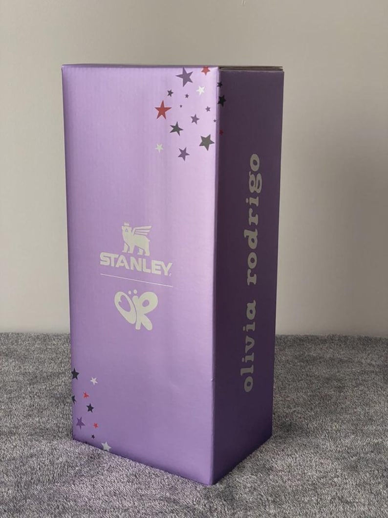 May include: A rectangular, lavender-colored box with the Stanley logo and the initials "OR" in white. The box is decorated with star shapes in various colors and the name "olivia rodrigo" is printed vertically on the side.