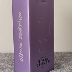 May include: A lavender box with the text "olivia rodrigo" in white, and a line drawing of a tumbler. The box also says "THE FLOWSTATE QUENCHER H2.0 TUMBLER" and indicates a 40 oz / 1.18L capacity.