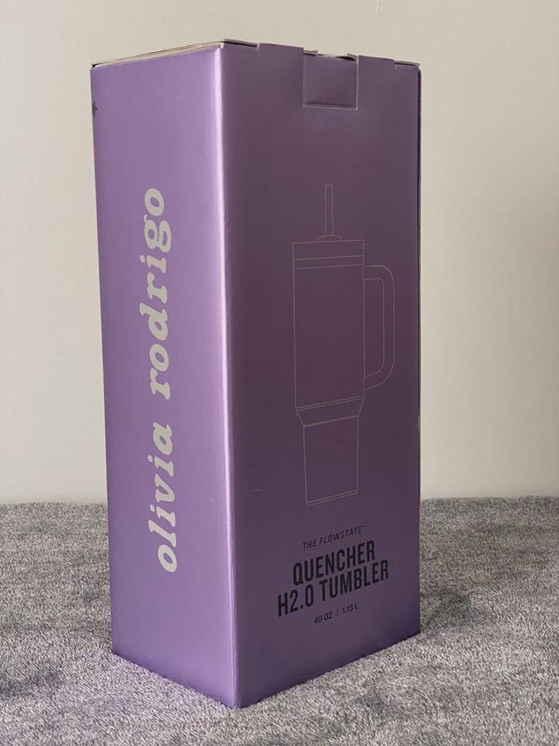 May include: A lavender box with the text "olivia rodrigo" in white, and a line drawing of a tumbler. The box also says "THE FLOWSTATE QUENCHER H2.0 TUMBLER" and indicates a 40 oz / 1.18L capacity.